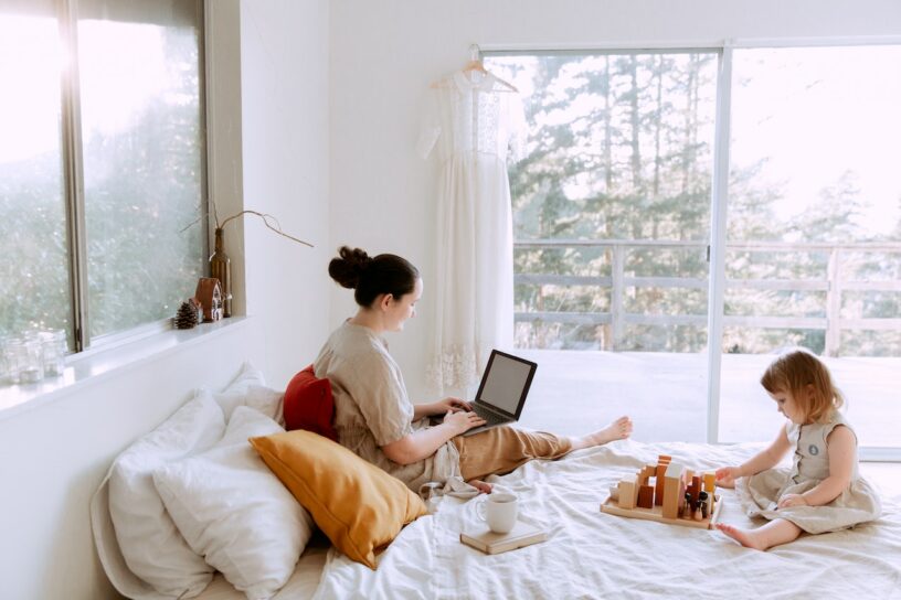 Adorable Girl Playing with Wooden Blocks, Sitting on the Bed, While Her Mother Laptops on a Sunny Morning