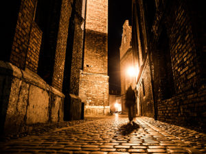 Illuminated cobbled street with light reflections on cobblestones in old historical city by night. Dark blurred silhouette of person evokes Jack the Ripper.